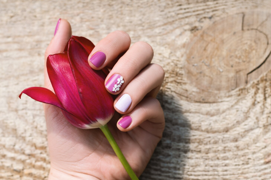 Hand with purple and pink nail art holding a red tulip against a wooden background, close-up view.
